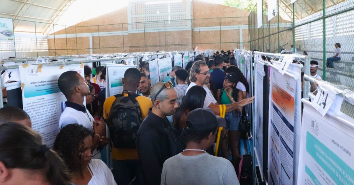 A imagem mostra um evento da Universidade Federal do Recôncavo da Bahia (UFRB) em um espaço coberto, onde várias pessoas estão reunidas em frente a painéis de apresentação. Os painéis parecem ser pôsteres acadêmicos e há muitas pessoas observando e discutindo o conteúdo.