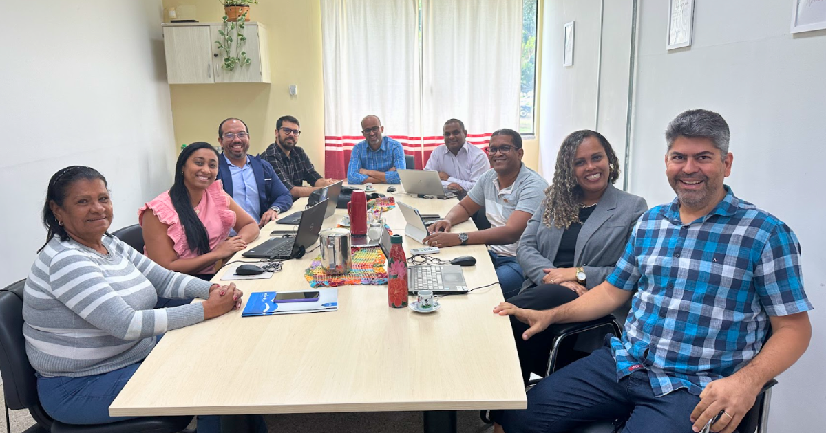 A imagem mostra um grupo de nove pessoas sentadas ao redor de uma mesa de reunião em um ambiente de escritório. A mesa é retangular e está cheia de laptops, garrafas de água, xícaras de café e alguns papéis. As pessoas estão sorrindo e parecem estar em um momento descontraído. Ao fundo, há uma janela com cortinas brancas e vermelhas que deixam entrar luz natural. Na parede à esquerda, há um armário suspenso com uma planta em cima. As pessoas estão vestidas de maneira casual e formal, com uma mistura de camisas, blusas e blazers.