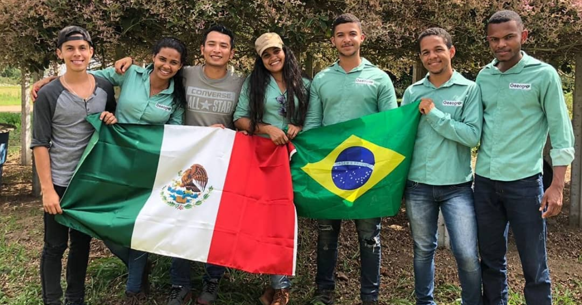 A imagem mostra um grupo de sete pessoas, todas sorrindo e posando para a foto. Eles estão ao ar livre, com árvores ao fundo. Duas pessoas no centro estão segurando bandeiras: a pessoa à esquerda segura a bandeira do México, que é verde, branca e vermelha com um brasão no centro, e a pessoa à direita segura a bandeira do Brasil, que é verde com um losango amarelo e um círculo azul com estrelas. A maioria das pessoas está vestindo camisas verdes claras com o logotipo "Geoparque" no peito. Uma pessoa à esquerda está usando uma camiseta cinza e um boné preto.